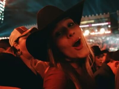 Mulher de chapeu de peao canta no meio do publico da Festa do Peao de Barretos, com a arena iluminada ao fundo