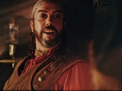 Tim Curry como o pirata Long John Silver, de barba e casaco vermelho, fala dentro de uma cabine de navio