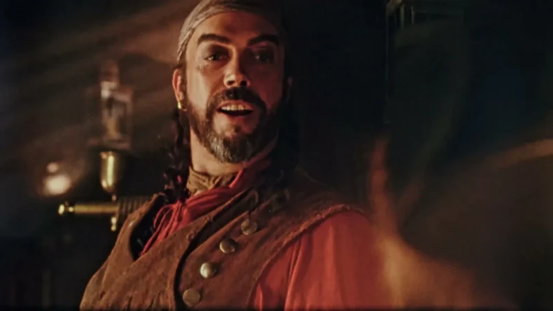 Tim Curry como o pirata Long John Silver, de barba e casaco vermelho, fala dentro de uma cabine de navio