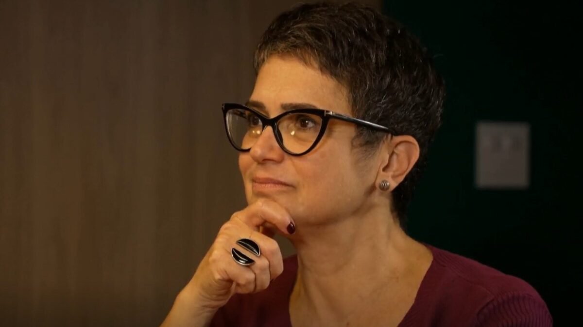 Sandra Annenberg de óculos, com a mão apoiada no queixo e expressão atenta, em blusa vinho