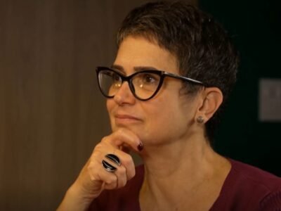 Sandra Annenberg de óculos, com a mão apoiada no queixo e expressão atenta, em blusa vinho