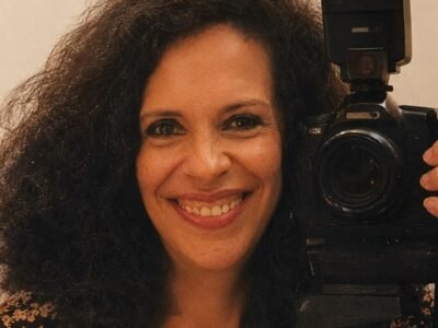 Gal Costa sorrindo, ao lado de uma camera fotografica sobre trive