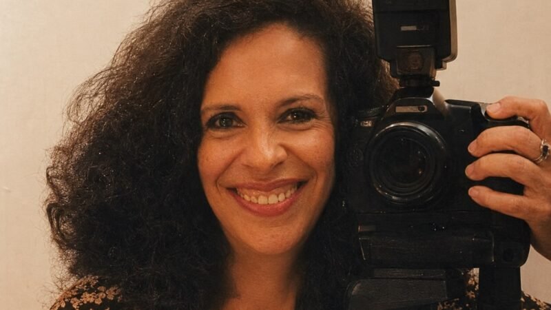 Gal Costa sorrindo, ao lado de uma camera fotografica sobre trive