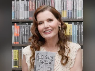 Geena Davis, de cabelo castanho comprido e blusa de tricô clara, sorri diante de uma estante de livros