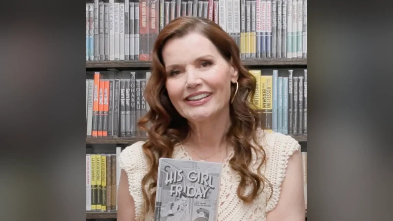 Geena Davis, de cabelo castanho comprido e blusa de tricô clara, sorri diante de uma estante de livros