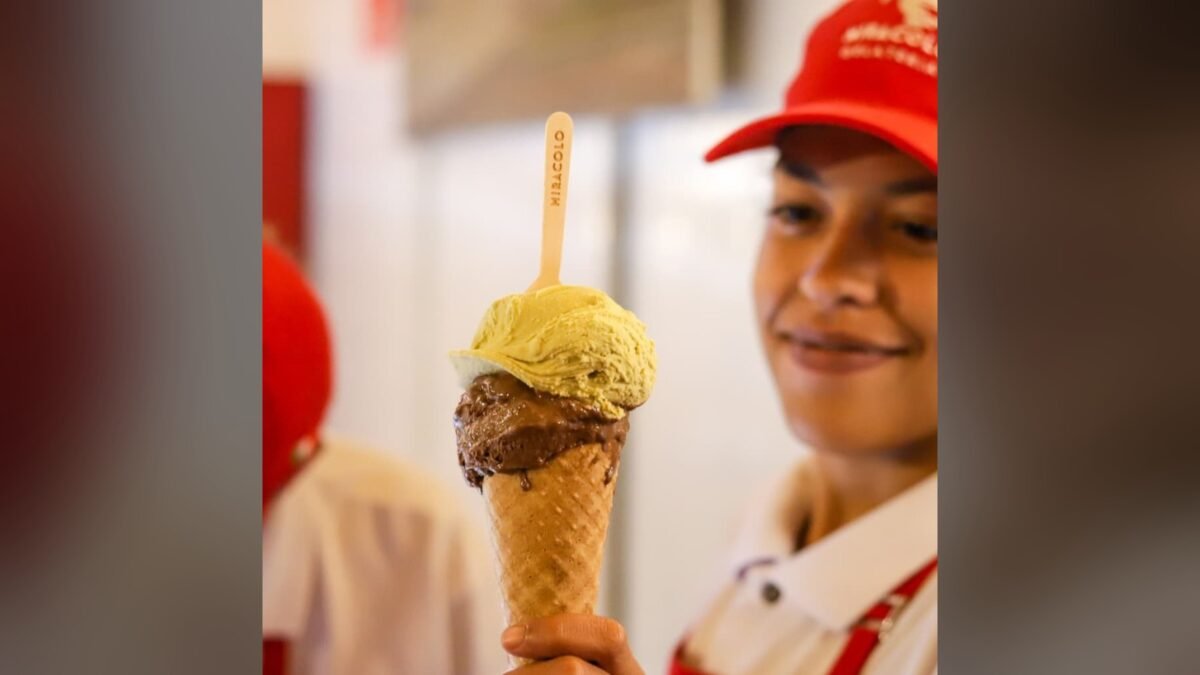 Atendente de boné vermelho da Gelateria Miracolo segura uma casquinha com gelato de chocolate e pistache