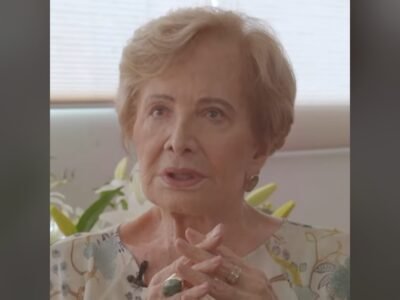 Gloria Menezes de blusa florida, com as maos juntas, em close durante entrevista em 2020