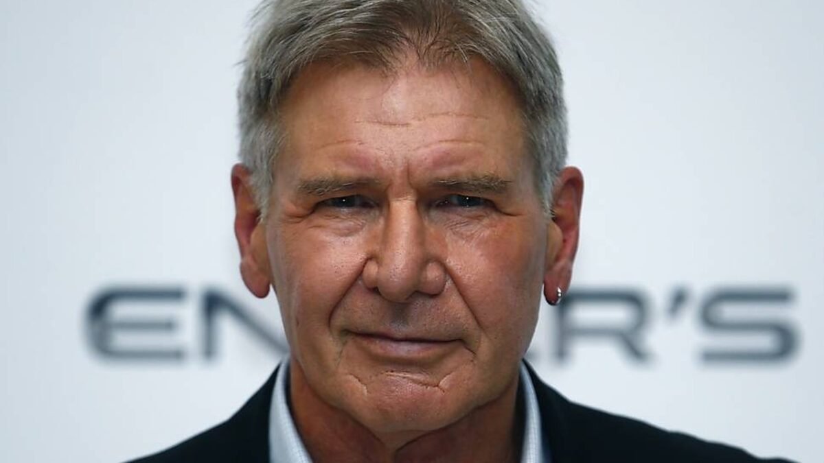 Harrison Ford em close, de terno escuro e brinco na orelha esquerda, diante de um painel de divulgacao
