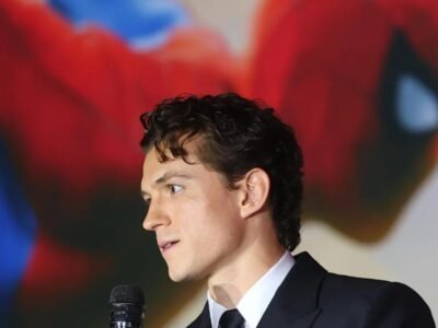 Tom Holland de terno fala à imprensa diante de uma arte do Homem-Aranha