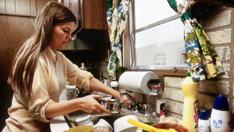 Mulher lavando louça na pia de uma cozinha de casa, de perfil, com a janela aberta ao lado