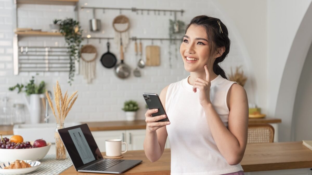 Mulher sorrindo com o celular na mão, de pé na cozinha de casa