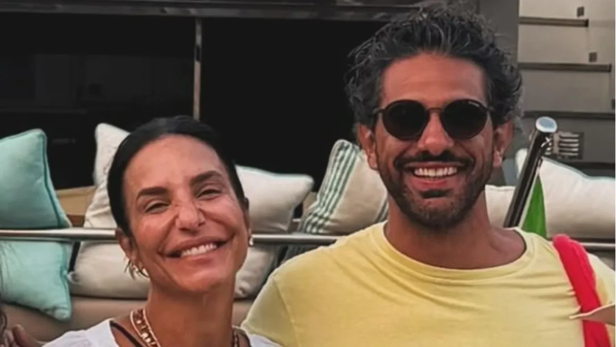Ivete Sangalo e Tiago Maia abraçados e sorrindo no deque de um barco, ele de camiseta amarela e óculos escuros