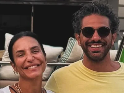 Ivete Sangalo e Tiago Maia abraçados e sorrindo no deque de um barco, ele de camiseta amarela e óculos escuros