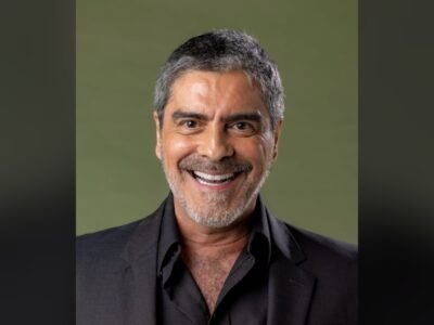 Junno Andrade sorri de camisa preta diante de um fundo verde-oliva, em retrato de estúdio