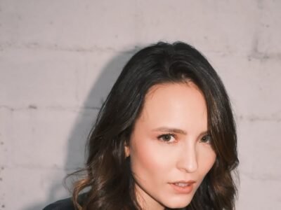Larissa Manoela de blazer escuro, olhando para a câmera, diante de uma parede de tijolo branco