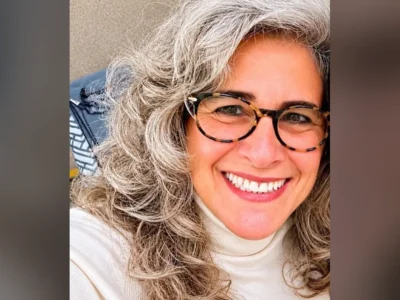 Lúcia Veríssimo, de cabelo grisalho e óculos de armação tartaruga, sorri de gola alta branca
