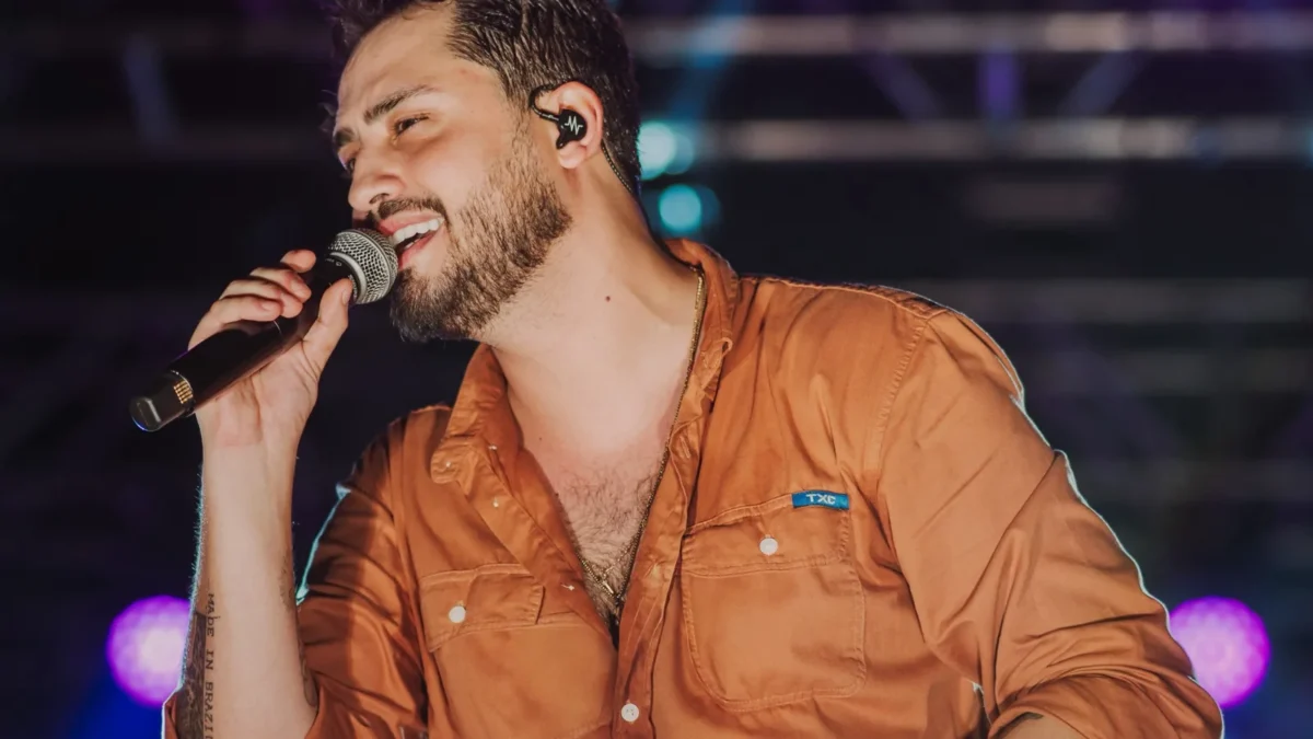 Matheus Vargas canta ao microfone em cima do palco, de camisa laranja, com luzes coloridas ao fundo
