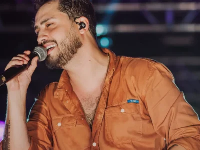 Matheus Vargas canta ao microfone em cima do palco, de camisa laranja, com luzes coloridas ao fundo