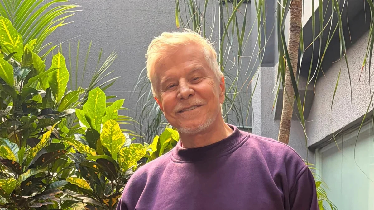 Miguel Falabella, de cabelo branco e moletom roxo, sorri em um jardim