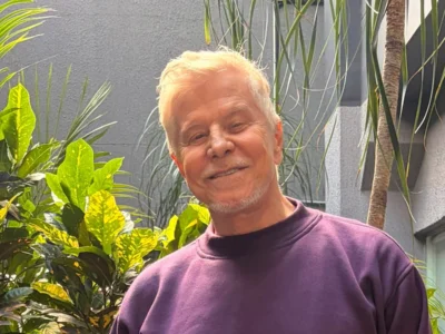 Miguel Falabella, de cabelo branco e moletom roxo, sorri em um jardim