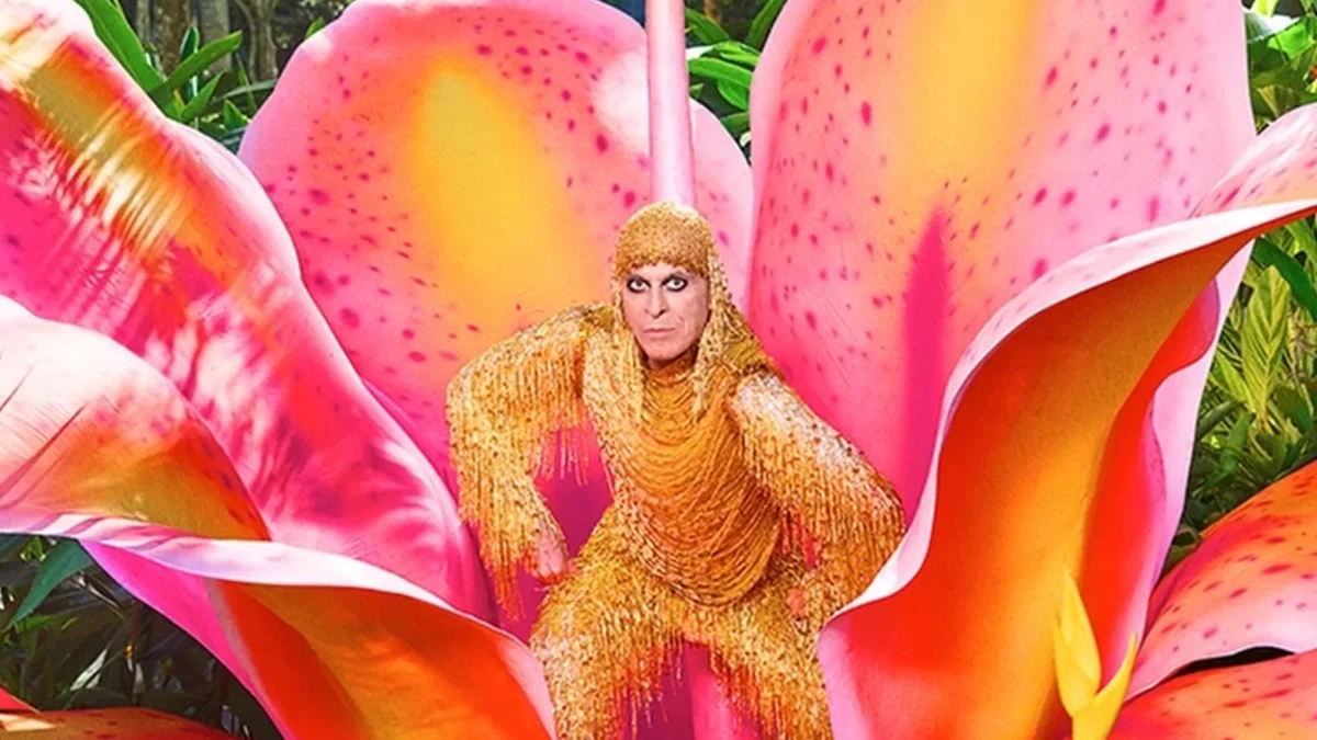 Ney Matogrosso de figurino dourado sentado no centro de uma flor rosa gigante, em foto da campanha da turnê
