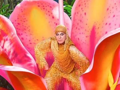 Ney Matogrosso de figurino dourado sentado no centro de uma flor rosa gigante, em foto da campanha da turnê