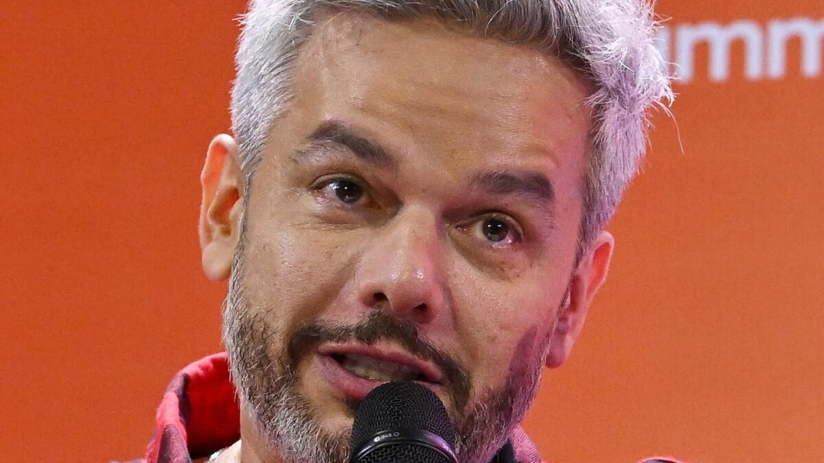 Otaviano Costa de camisa xadrez vermelha, cabelo grisalho, falando ao microfone