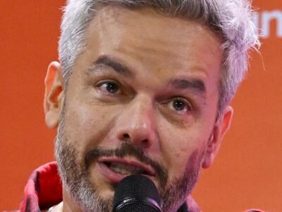 Otaviano Costa de camisa xadrez vermelha, cabelo grisalho, falando ao microfone