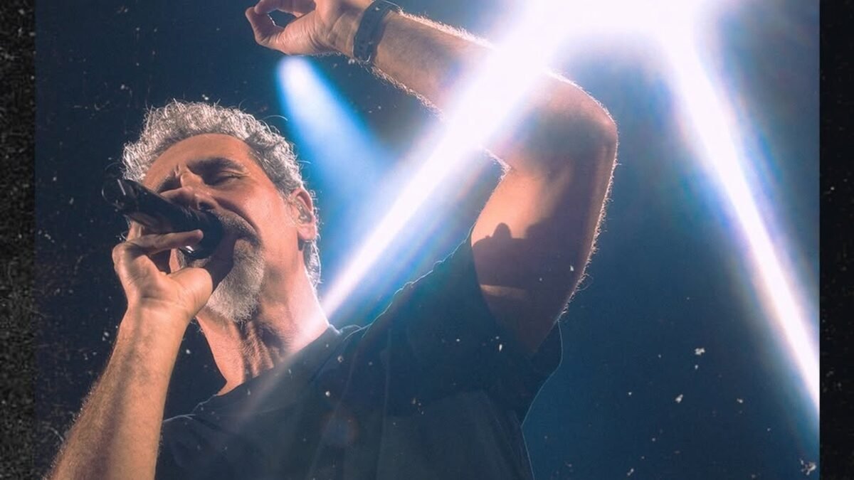 Cantor do System of a Down canta ao microfone sob um facho de luz branca no palco