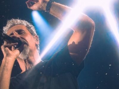 Cantor do System of a Down canta ao microfone sob um facho de luz branca no palco