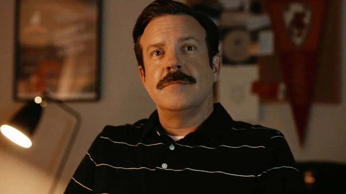 Jason Sudeikis como Ted Lasso, de bigode e camisa polo escura, sentado num escritório