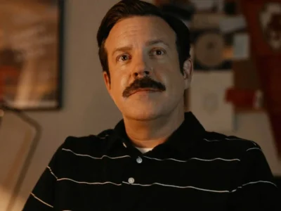Jason Sudeikis como Ted Lasso, de bigode e camisa polo escura, sentado num escritório