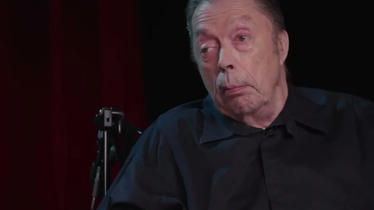 Tim Curry, de camisa preta e cabelo grisalho, olha de lado durante entrevista com fundo escuro