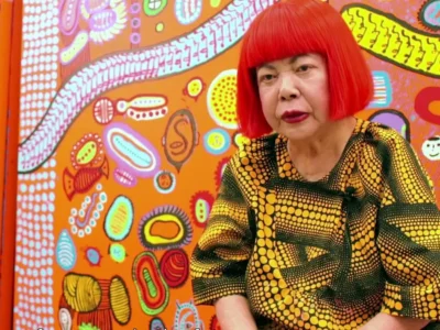 Yayoi Kusama, de peruca vermelha e roupa de bolinhas, sentada diante de uma pintura colorida dela