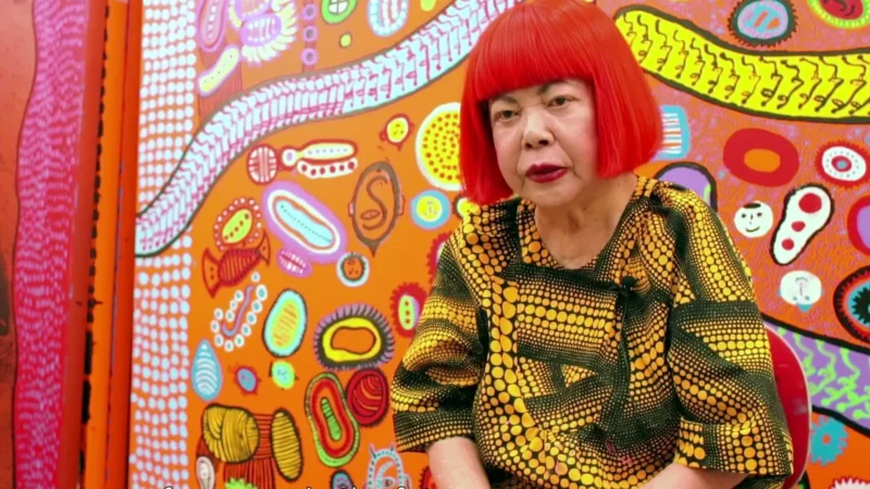 Yayoi Kusama, de peruca vermelha e roupa de bolinhas, sentada diante de uma pintura colorida dela