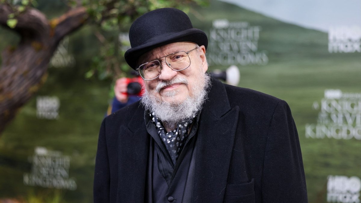 George R.R. Martin de boina e casaco preto em evento ao ar livre.