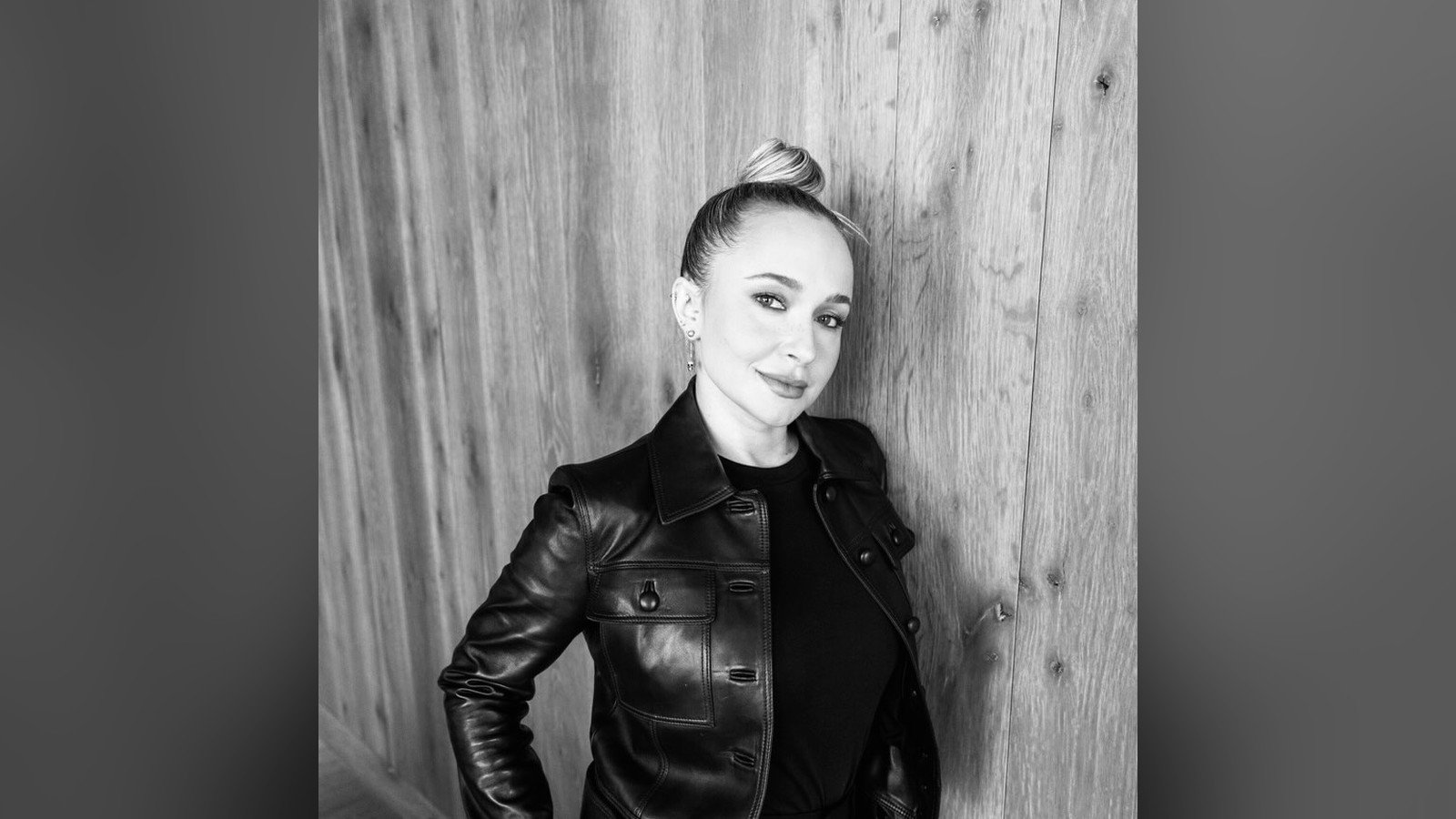 Hayden Panettiere em foto preto e branco, de jaqueta de couro, encostada em uma parede de madeira