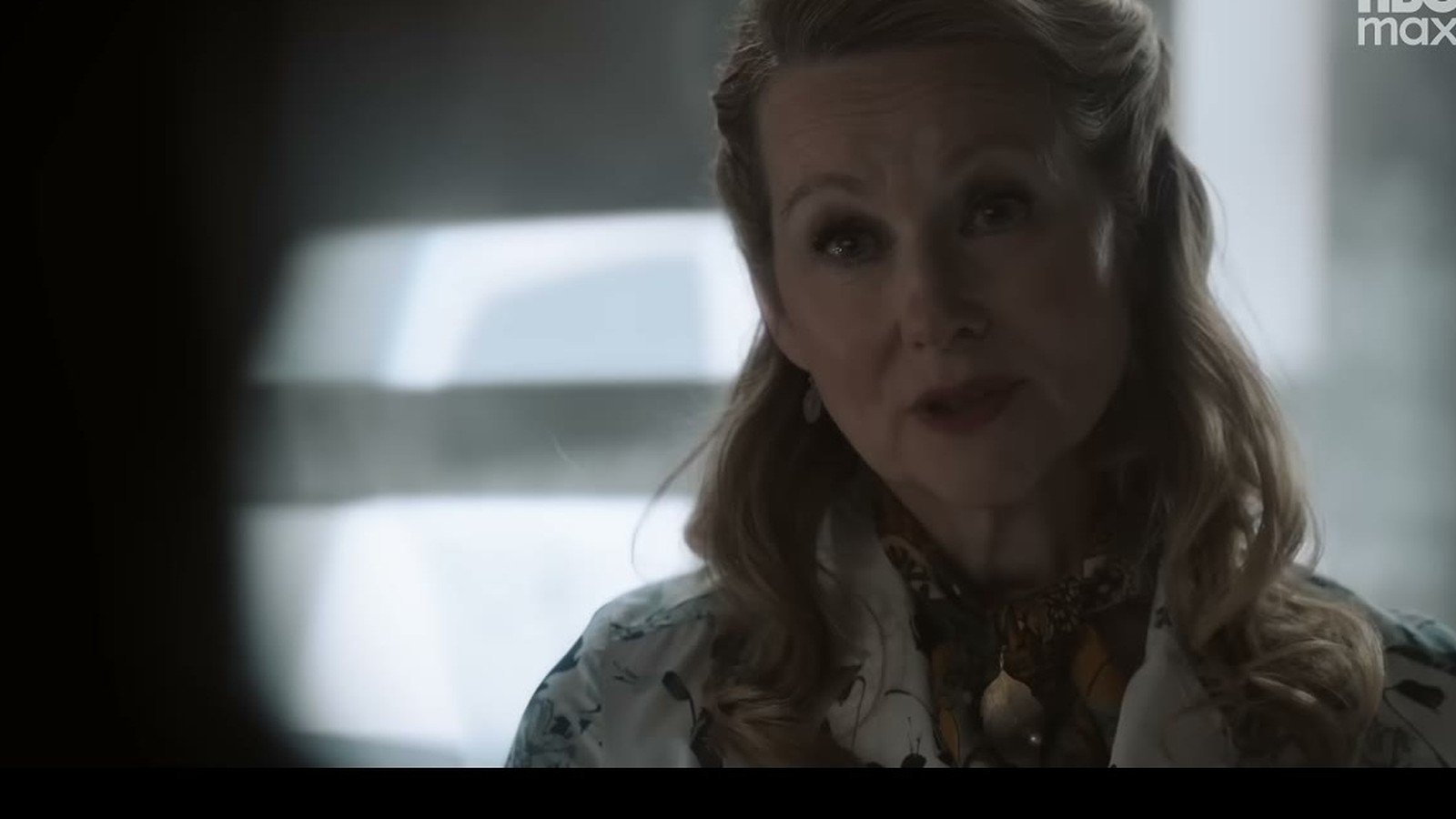 Atriz loira de blusa estampada fala diante de uma janela iluminada numa cena da serie