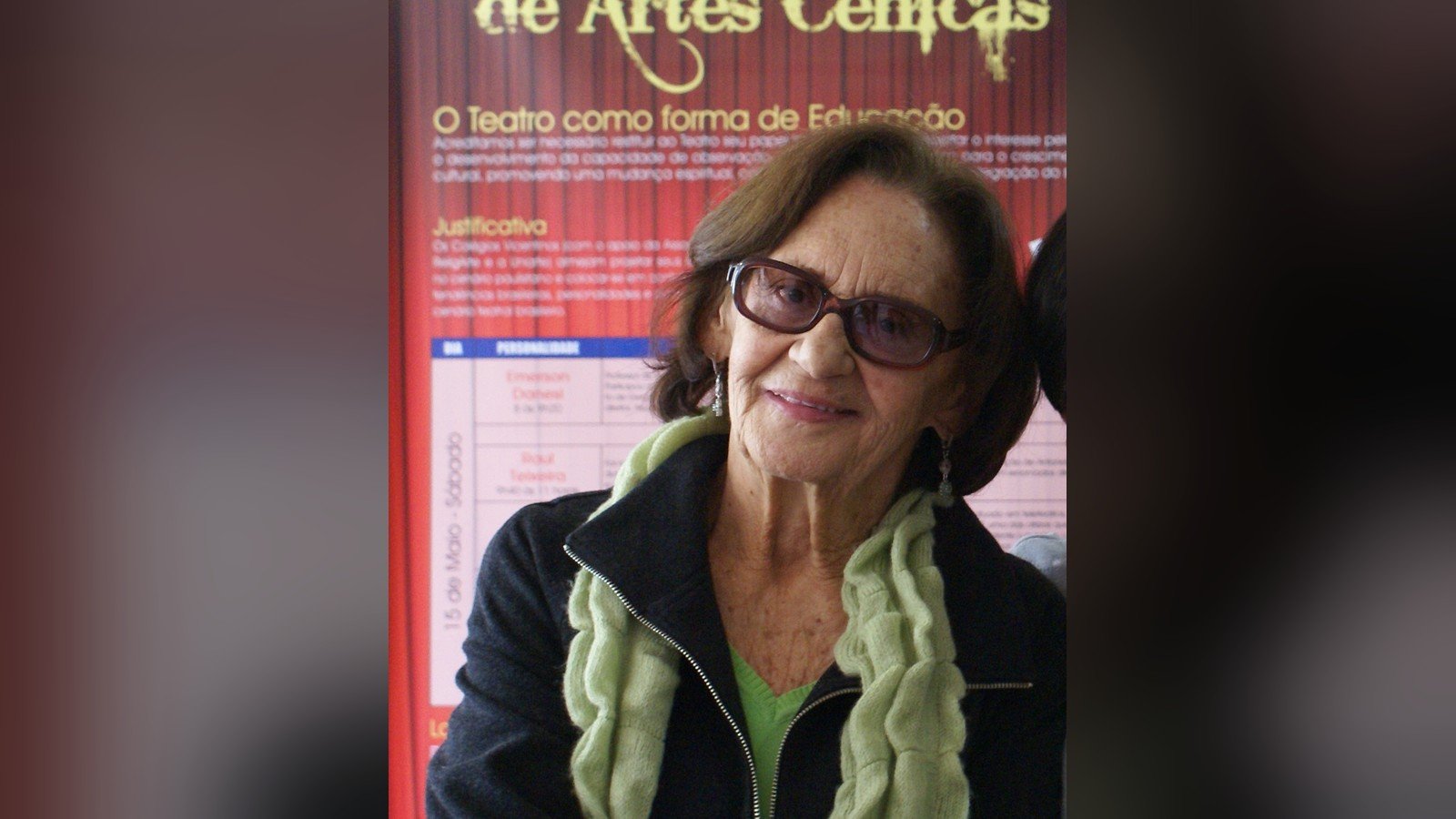 Laura Cardoso de oculos escuros e casaco preto diante de um painel de evento de artes cenicas
