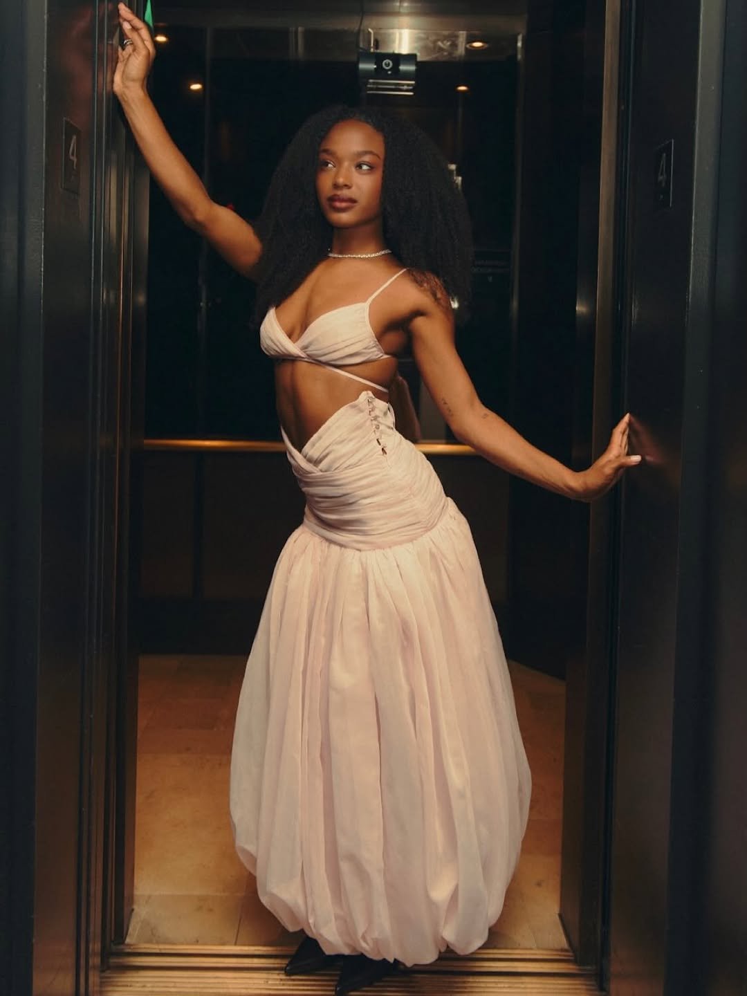 Maya Boyd de vestido claro longo, em pé no vao de uma porta