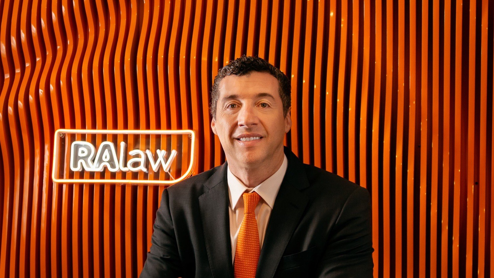 O advogado Marcelo Robalinho, de terno escuro e gravata laranja, diante do letreiro de neon do escritório RA Law