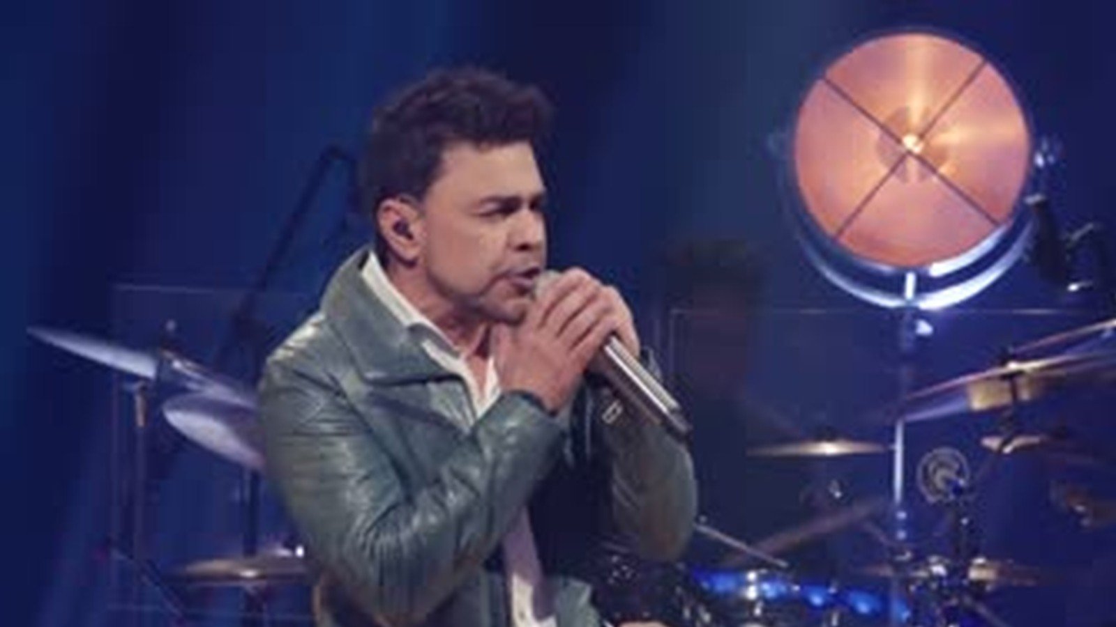Zezé Di Camargo de jaqueta clara canta ao microfone no palco, com a bateria da banda ao fundo