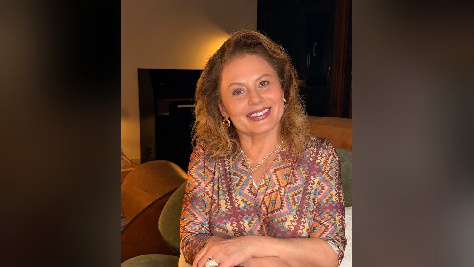Vera Fischer sentada num sofá, de vestido estampado, sorrindo para a câmera