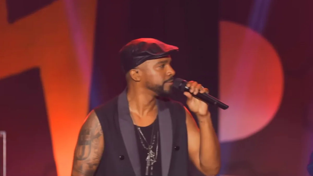 Alexandre Pires, de boina e colete preto, canta ao microfone num palco com luz vermelha
