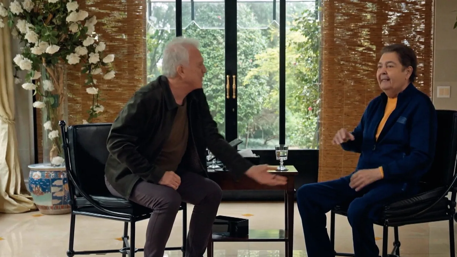 Faustão, de agasalho azul e camiseta amarela, conversa com Pedro Bial sentados frente a frente em uma sala