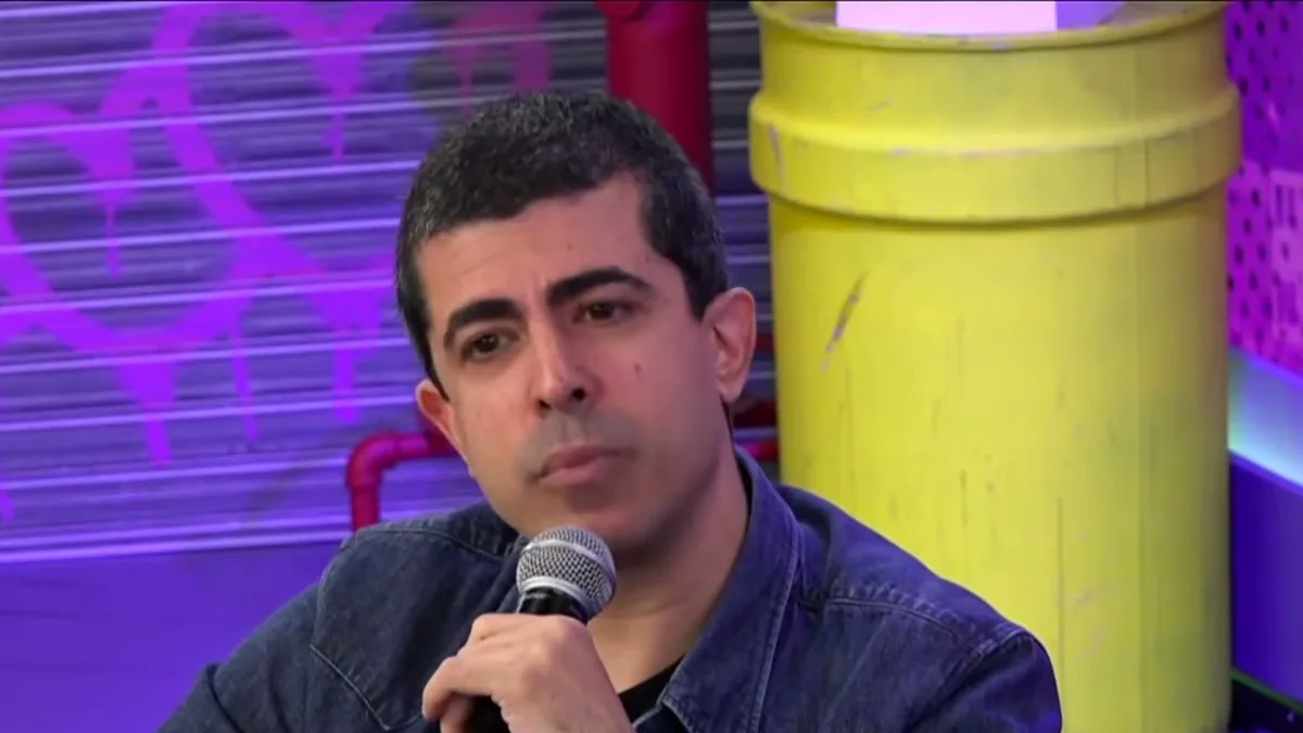 Marcius Melhem, de jaqueta jeans, segurando o microfone num estúdio de TV com parede roxa