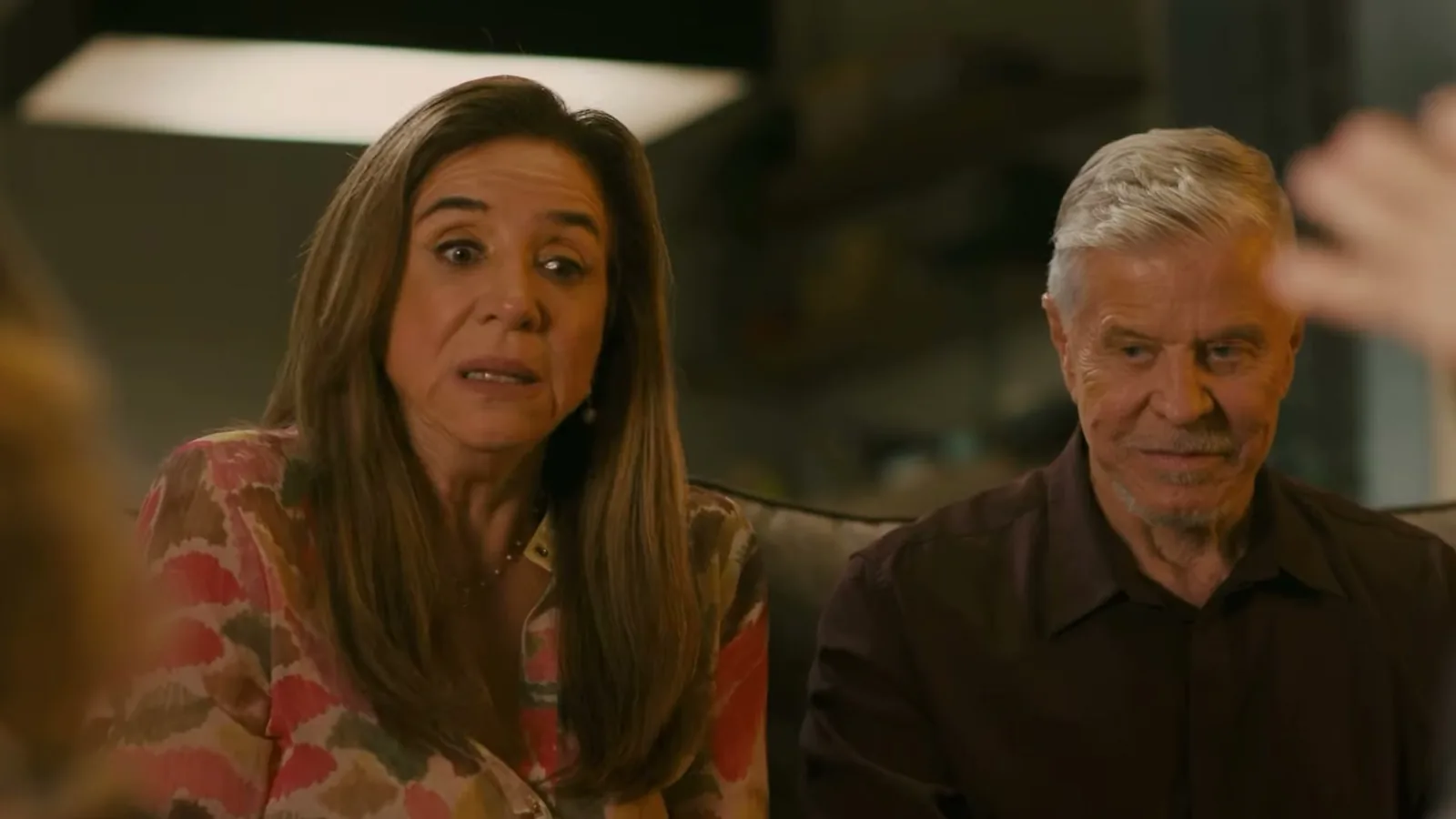 Marisa Orth e Miguel Falabella sentados lado a lado em um sofá, em cena do filme O Pecado Mora em Cima