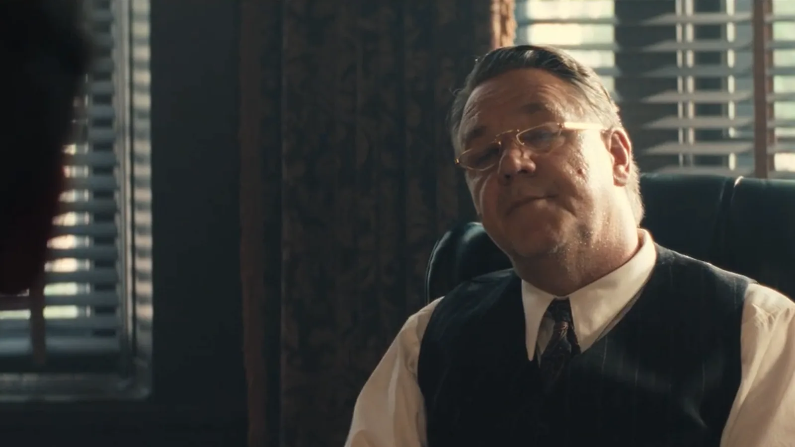 Russell Crowe, de óculos, colete e gravata, sentado atrás de uma mesa num escritório, no papel do professor Henry Murray