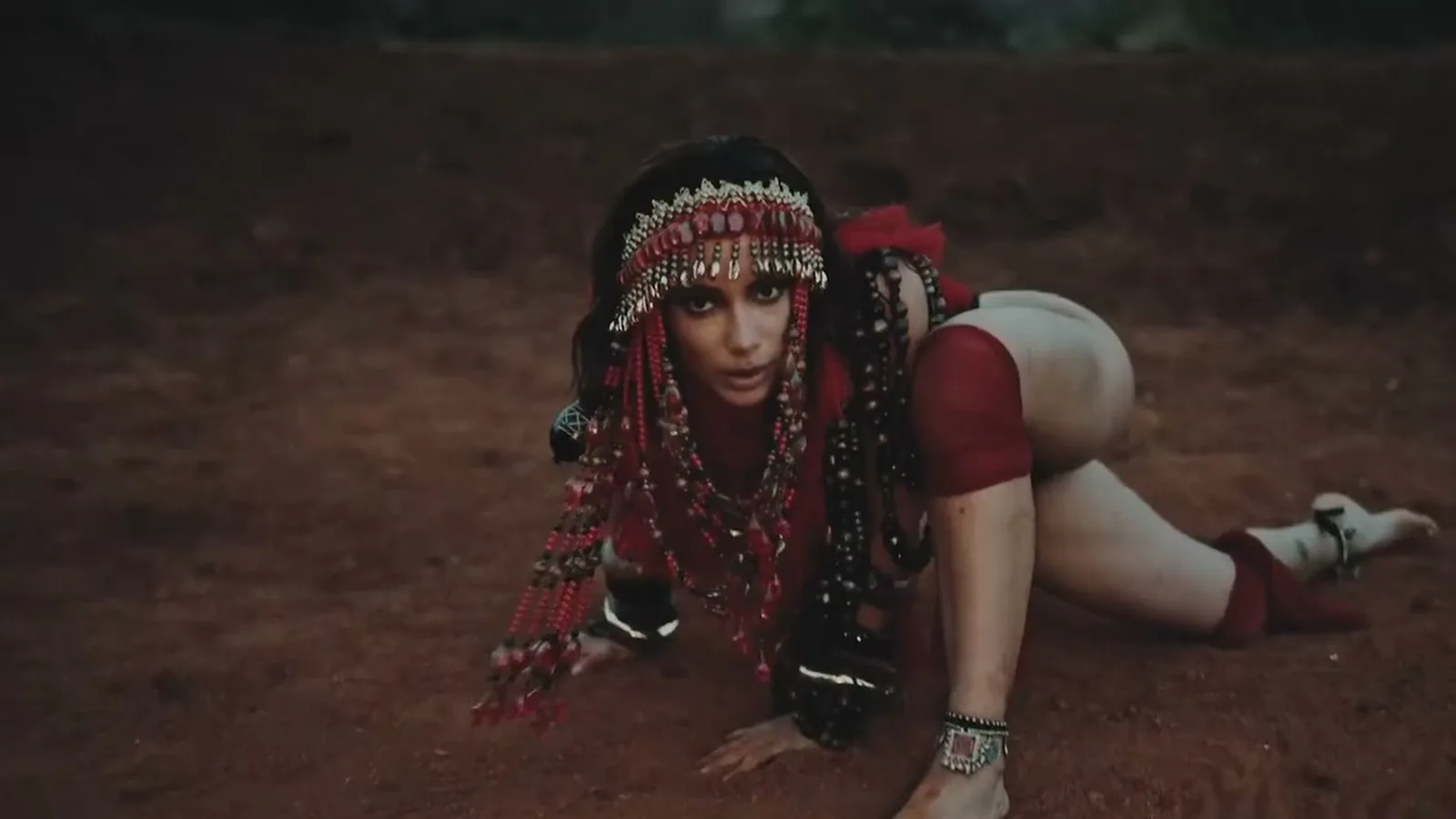Anitta com adereço de contas vermelhas na cabeça, apoiada no chão de terra, no clipe de Choka Choka
