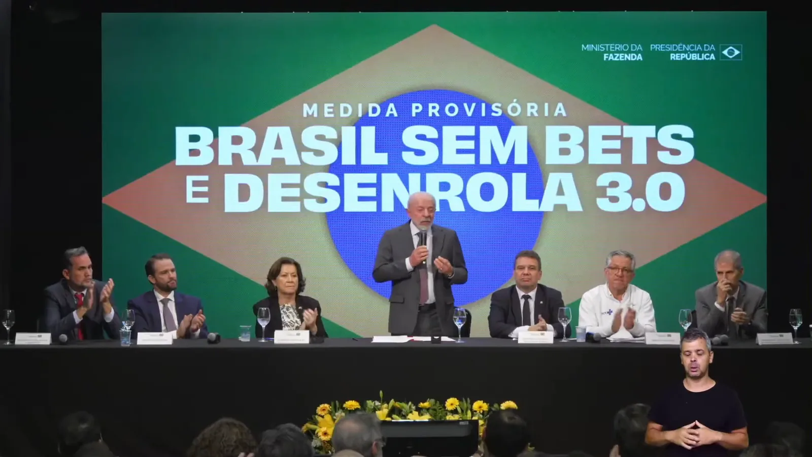 Lula discursa em pé diante de um painel verde e amarelo com os dizeres Medida Provisória Brasil sem bets e Desenrola 3.0, ao lado de ministros sentados à mesa